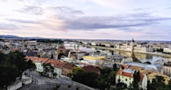 A panoramic view of a cityscape with a river running through it. The architecture is diverse with buildings of various styles and colors, including a prominent parliament building by the water. The sky is mostly cloudy with hints of pink and blue.