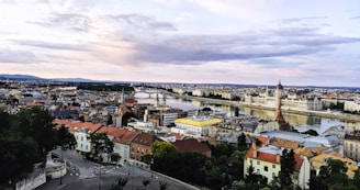 A panoramic view of a cityscape with a river running through it. The architecture is diverse with buildings of various styles and colors, including a prominent parliament building by the water. The sky is mostly cloudy with hints of pink and blue.