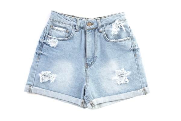 A pair of light blue distressed denim shorts with frayed details and rolled cuffs. The front has two pockets and a waistband with a metal button.