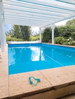 An outdoor swimming pool is surrounded by a stone deck, with a pair of swimming goggles placed on the pool's edge. The pool is beneath a white, open pergola, and lush green trees and bushes are visible in the background.