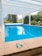 An outdoor swimming pool is surrounded by a stone deck, with a pair of swimming goggles placed on the pool's edge. The pool is beneath a white, open pergola, and lush green trees and bushes are visible in the background.