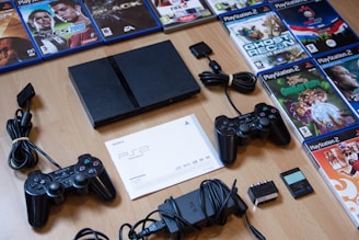 A collection of PlayStation 2 gaming items spread out on a wooden surface includes the console, multiple game discs with cases arranged in a semi-circle, two black controllers, a memory card, various cables, and an instruction manual.