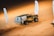 A rugged off-road vehicle is driving across sand dunes, kicking up a significant amount of dust. The vehicle is adorned with various sponsor logos and has hefty tires suitable for sandy terrain. Nearby, several banners or flags with Arabic writing are planted in the sand, swaying gently in the breeze.