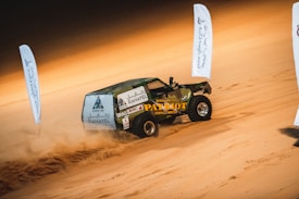 A rugged off-road vehicle is driving across sand dunes, kicking up a significant amount of dust. The vehicle is adorned with various sponsor logos and has hefty tires suitable for sandy terrain. Nearby, several banners or flags with Arabic writing are planted in the sand, swaying gently in the breeze.