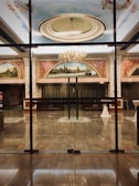 A luxurious and elegant interior space with a polished marble floor, reflecting the light from a grand chandelier. There are large painted murals above the entrance, framed with ornate moldings. Glass doors separate the hallway from what appears to be a ballroom or large reception area. Tables are set with white tablecloths, seemingly prepared for an event.