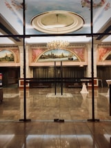 A luxurious and elegant interior space with a polished marble floor, reflecting the light from a grand chandelier. There are large painted murals above the entrance, framed with ornate moldings. Glass doors separate the hallway from what appears to be a ballroom or large reception area. Tables are set with white tablecloths, seemingly prepared for an event.