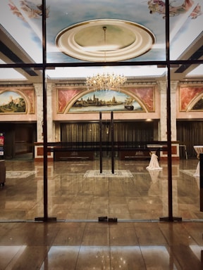 A luxurious and elegant interior space with a polished marble floor, reflecting the light from a grand chandelier. There are large painted murals above the entrance, framed with ornate moldings. Glass doors separate the hallway from what appears to be a ballroom or large reception area. Tables are set with white tablecloths, seemingly prepared for an event.