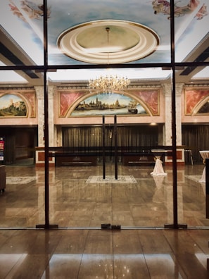 A luxurious and elegant interior space with a polished marble floor, reflecting the light from a grand chandelier. There are large painted murals above the entrance, framed with ornate moldings. Glass doors separate the hallway from what appears to be a ballroom or large reception area. Tables are set with white tablecloths, seemingly prepared for an event.
