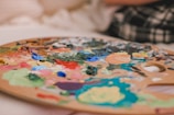 A wooden painter's palette covered in vibrant dabs of various colored paints, including blue, green, orange, and red, with a blurred background.