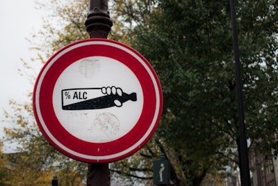 A round traffic sign with a red border against a backdrop of trees and poles. Inside the sign is an image of a hand holding a bottle with the text '% ALC' printed on it.