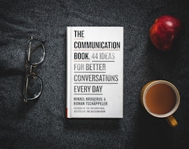 A book titled 'The Communication Book: 44 Ideas for Better Conversations Every Day' is placed on a dark textured surface. Next to it are a pair of glasses, an apple, and a cup of coffee.