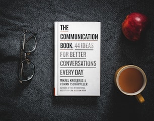 A book titled 'The Communication Book: 44 Ideas for Better Conversations Every Day' is placed on a dark textured surface. Next to it are a pair of glasses, an apple, and a cup of coffee.