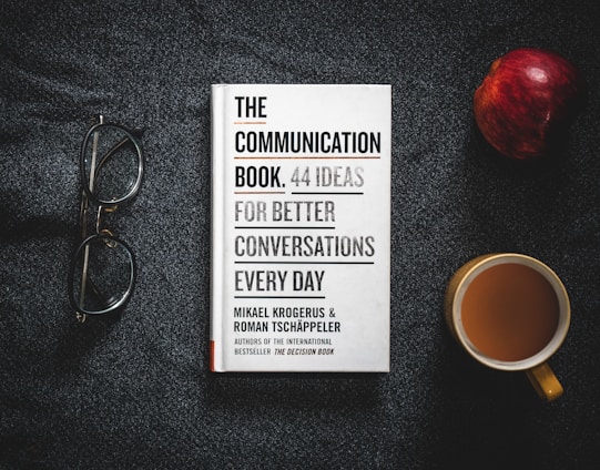 A book titled 'The Communication Book: 44 Ideas for Better Conversations Every Day' is placed on a dark textured surface. Next to it are a pair of glasses, an apple, and a cup of coffee.