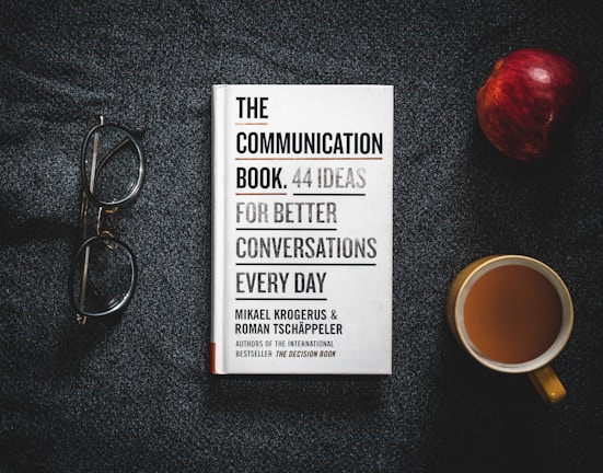 A book titled 'The Communication Book: 44 Ideas for Better Conversations Every Day' is placed on a dark textured surface. Next to it are a pair of glasses, an apple, and a cup of coffee.