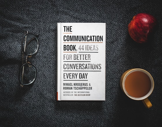 A book titled 'The Communication Book: 44 Ideas for Better Conversations Every Day' is placed on a dark textured surface. Next to it are a pair of glasses, an apple, and a cup of coffee.