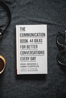 A book titled 'The Communication Book. 44 Ideas for Better Conversations Every Day' by Mikael Krogerus and Roman Tschäppeler is placed on a dark textured surface. There are headphones to the right and a cup on the left.