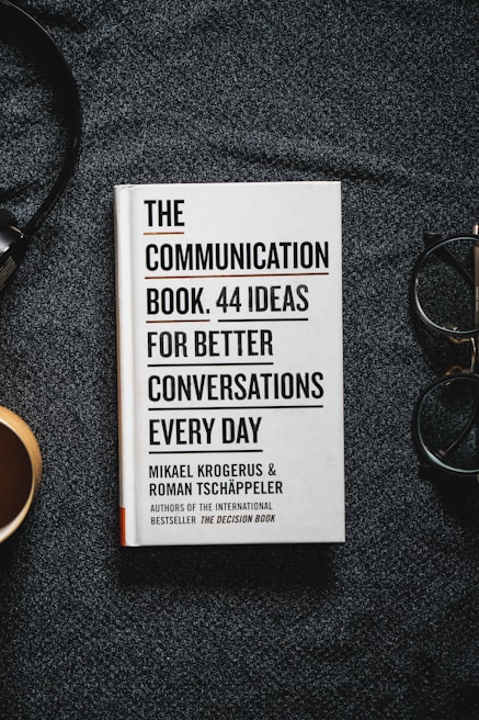 A book titled 'The Communication Book. 44 Ideas for Better Conversations Every Day' by Mikael Krogerus and Roman Tschäppeler is placed on a dark textured surface. There are headphones to the right and a cup on the left.