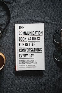 A book titled 'The Communication Book. 44 Ideas for Better Conversations Every Day' by Mikael Krogerus and Roman Tschäppeler is placed on a dark textured surface. There are headphones to the right and a cup on the left.