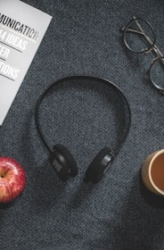 A pair of black headphones is placed on a textured dark gray surface, surrounded by an apple, a brown coffee cup, glasses, and a magazine. The magazine has a visible text featuring the word 'COMMUNICATION' prominently.