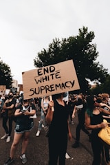 A group of people are participating in a protest, with one individual prominently holding a cardboard sign reading 'END WHITE SUPREMACY'. The participants are wearing casual clothing, and some have face coverings. The setting appears to be outdoors with trees in the background.