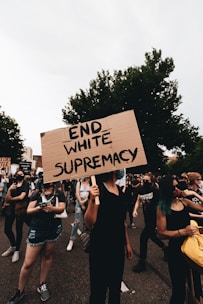 A group of people are participating in a protest, with one individual prominently holding a cardboard sign reading 'END WHITE SUPREMACY'. The participants are wearing casual clothing, and some have face coverings. The setting appears to be outdoors with trees in the background.