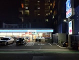 A convenience store is illuminated at night, with two cars parked in front. The store's bright interior contrasts with the surrounding darkness. A sign and a bicycle are visible on the right side.
