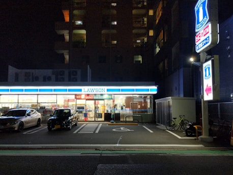 A convenience store is illuminated at night, with two cars parked in front. The store's bright interior contrasts with the surrounding darkness. A sign and a bicycle are visible on the right side.
