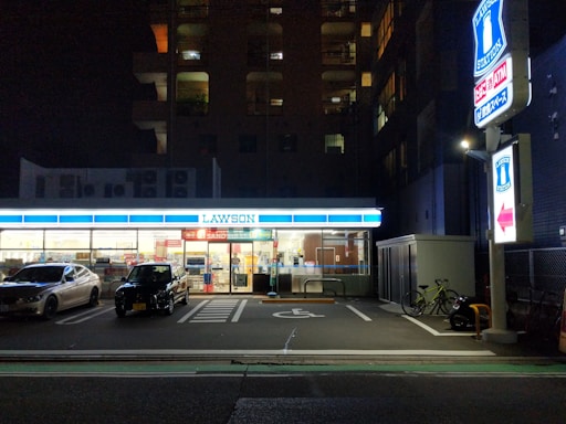 A convenience store is illuminated at night, with two cars parked in front. The store's bright interior contrasts with the surrounding darkness. A sign and a bicycle are visible on the right side.