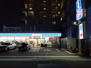 A convenience store is illuminated at night, with two cars parked in front. The store's bright interior contrasts with the surrounding darkness. A sign and a bicycle are visible on the right side.