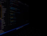 A close-up view of a computer screen displaying code in a dark setting. The code appears to be written in a text editor with syntax highlighting, featuring various lines of text and numbers. The surroundings are dimly lit, emphasizing the bright screen against the dark background.