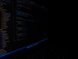 A close-up view of a computer screen displaying code in a dark setting. The code appears to be written in a text editor with syntax highlighting, featuring various lines of text and numbers. The surroundings are dimly lit, emphasizing the bright screen against the dark background.