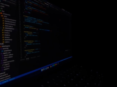 A close-up view of a computer screen displaying code in a dark setting. The code appears to be written in a text editor with syntax highlighting, featuring various lines of text and numbers. The surroundings are dimly lit, emphasizing the bright screen against the dark background.