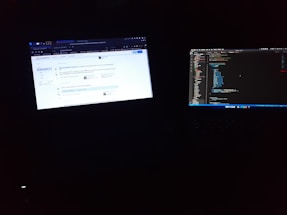 Two laptop screens are illuminated in a dark environment. One screen displays a web browser with a question on a coding forum, while the other screen shows a code editor with lines of code.