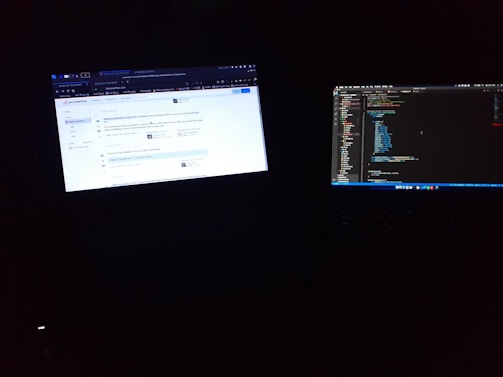 Two laptop screens are illuminated in a dark environment. One screen displays a web browser with a question on a coding forum, while the other screen shows a code editor with lines of code.