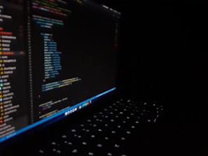 A dark image featuring a laptop with a visible keyboard illuminated by screen light. The screen displays multicolored code in an editor, showing lines of programming in different text colors.