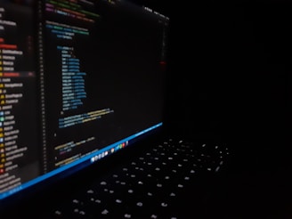 A dark image featuring a laptop with a visible keyboard illuminated by screen light. The screen displays multicolored code in an editor, showing lines of programming in different text colors.