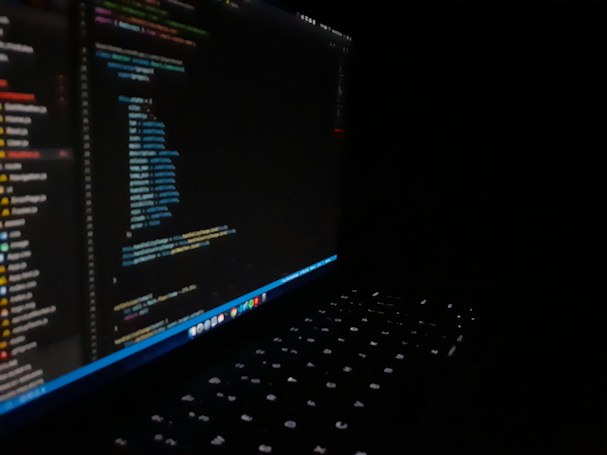 A dark image featuring a laptop with a visible keyboard illuminated by screen light. The screen displays multicolored code in an editor, showing lines of programming in different text colors.