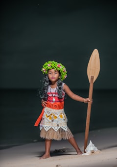 A young child is dressed in a colorful costume reminiscent of Polynesian attire, standing confidently on a sandy beach. The outfit includes a grass skirt, patterned top, and a red sash. A leafy flower crown adorns the child's head, and a wooden paddle is held upright. The background features a dark, blurred seascape.
