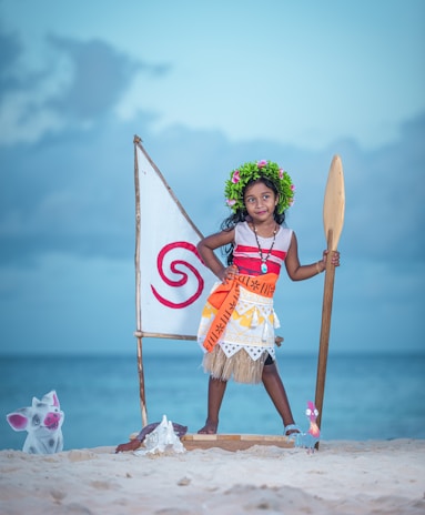 A child is dressed in a tropical-themed costume, standing on a sandy beach with an ocean backdrop. The costume includes a colorful skirt, a sash, and a floral headpiece. The child holds a wooden paddle next to a decorative sail with a red spiral symbol. There are cartoon cutouts of a pig and a rooster near the child's feet.