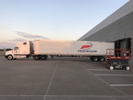 A large white semi-truck is parked outside a warehouse loading dock, positioned to unload or load goods. The truck features a logo with the word 'Innovativos' on the side. Nearby, there is a red and black mechanical lift labeled 'SkyJack' on the ground. The setting is an open area with clear skies in the background.