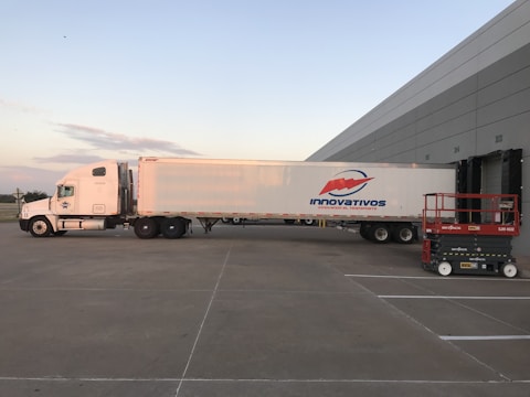 A large white semi-truck is parked outside a warehouse loading dock, positioned to unload or load goods. The truck features a logo with the word 'Innovativos' on the side. Nearby, there is a red and black mechanical lift labeled 'SkyJack' on the ground. The setting is an open area with clear skies in the background.