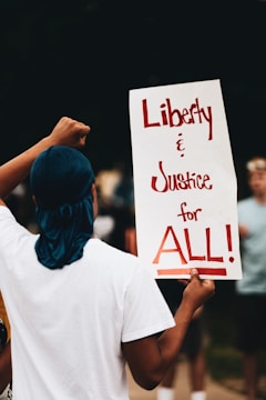 A person wearing a white shirt and blue headscarf holds up a protest sign with the words 'Liberty & Justice for ALL!' written in bold red letters. The background shows a blurred crowd, indicating a demonstration or rally setting.