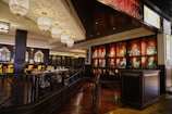 An elegantly designed restaurant interior featuring intricate chandeliers that cast soft lighting, with dark wooden paneling and artistic displays along the walls. The area has neatly arranged tables with dining settings, and high-backed chairs against a backdrop of stylish decor with Asian-inspired wall patterns.
