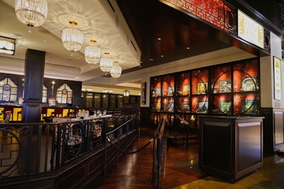 An elegantly designed restaurant interior featuring intricate chandeliers that cast soft lighting, with dark wooden paneling and artistic displays along the walls. The area has neatly arranged tables with dining settings, and high-backed chairs against a backdrop of stylish decor with Asian-inspired wall patterns.