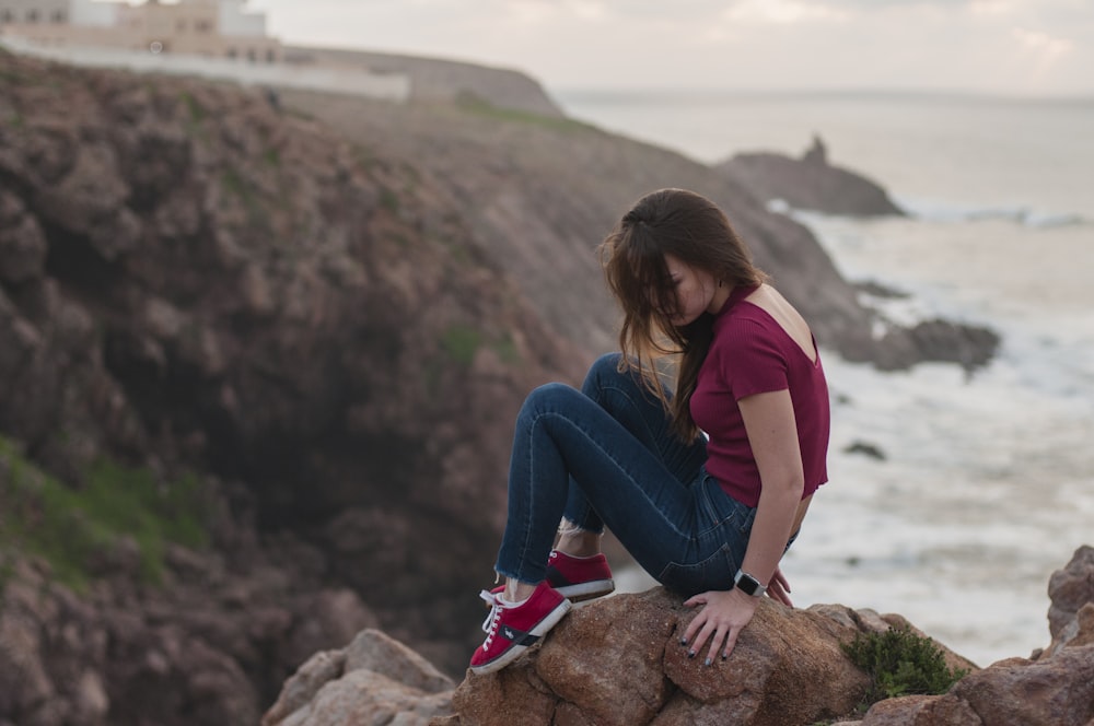 woman in pink shirt and blue denim jeans sitting on brown rock during daytime