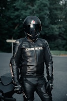 A person wearing a full black motorcycle suit with a matching helmet stands outdoors. The helmet is glossy with a logo, and the suit has the brand name printed on it. The background consists of blurred green foliage and a road surface.