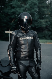 A person wearing a full black motorcycle suit with a matching helmet stands outdoors. The helmet is glossy with a logo, and the suit has the brand name printed on it. The background consists of blurred green foliage and a road surface.