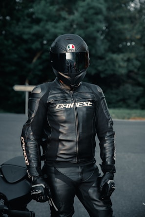 A person wearing a full black motorcycle suit with a matching helmet stands outdoors. The helmet is glossy with a logo, and the suit has the brand name printed on it. The background consists of blurred green foliage and a road surface.