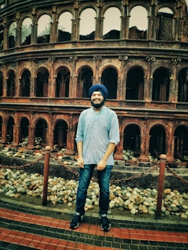 A man wearing a turban and casual clothing stands in front of a historical structure resembling an ancient amphitheater. The foreground features a path made of red and dark gray bricks, bordered by pebbles and chain fencing.