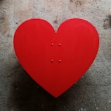 A large red heart shape placed on a textured concrete background. The heart is symmetrical and vibrant, standing out against the muted and rough surface.